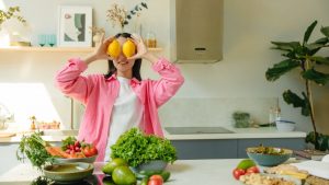 A woman playfully holding lemons over her eyes in a bright, lively kitchen setting filled with fresh vegetables.